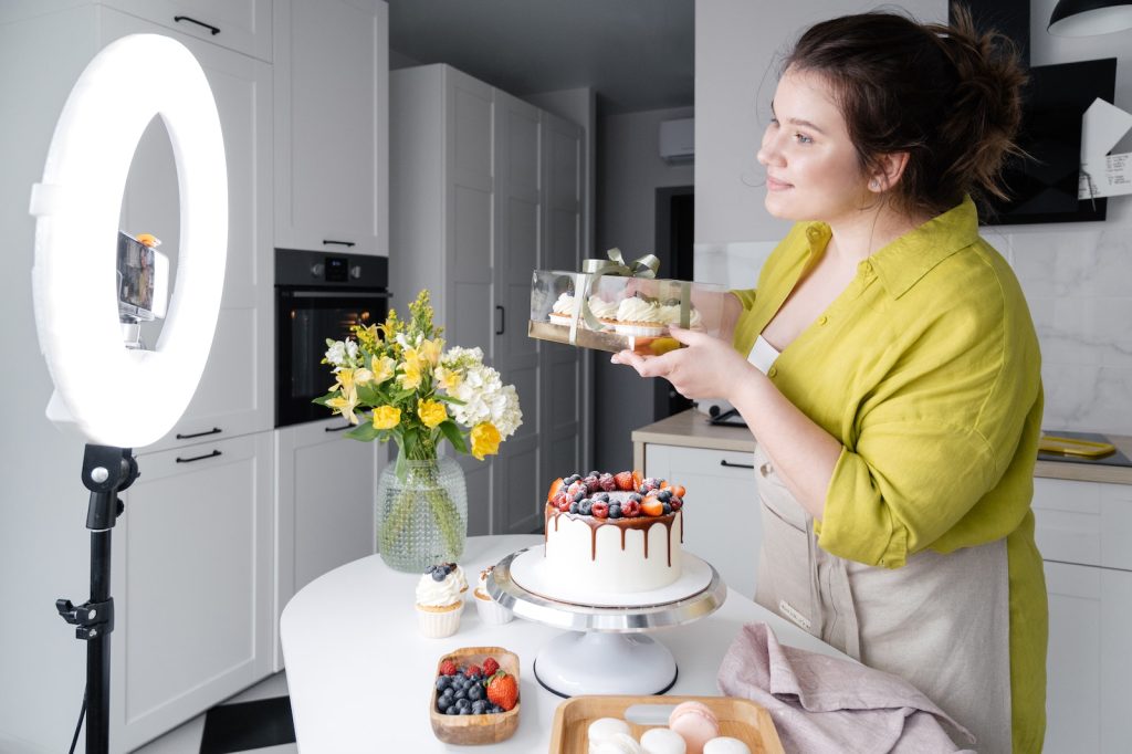 Side view of positive young female food blogger in casual clothes and apron introducing yummy cakes and smiling while standing in front of ring lamp and recording vlog on smartphone